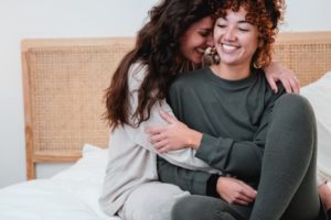 Ein lesbisches Paar sitzt auf dem Bett, lacht und umarmt sich.